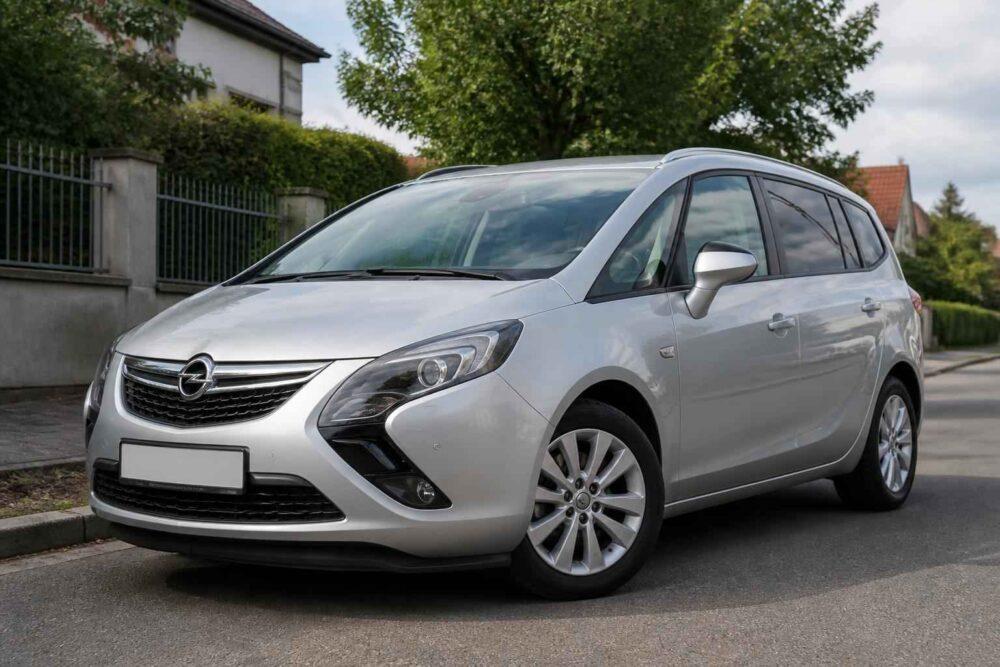 Srebrny Opel Zafira zaparkowany przy ulicy przed domem z ogrodem